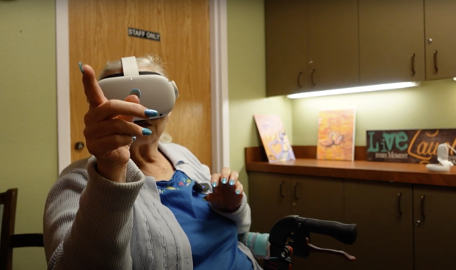 An older woman wearing a white virtual reality headset sits in a chair, extending one hand forward with her fingers pointing, as if interacting with a virtual object. She has vibrant, teal-painted nails and is dressed in a blue shirt with a light gray cardigan.